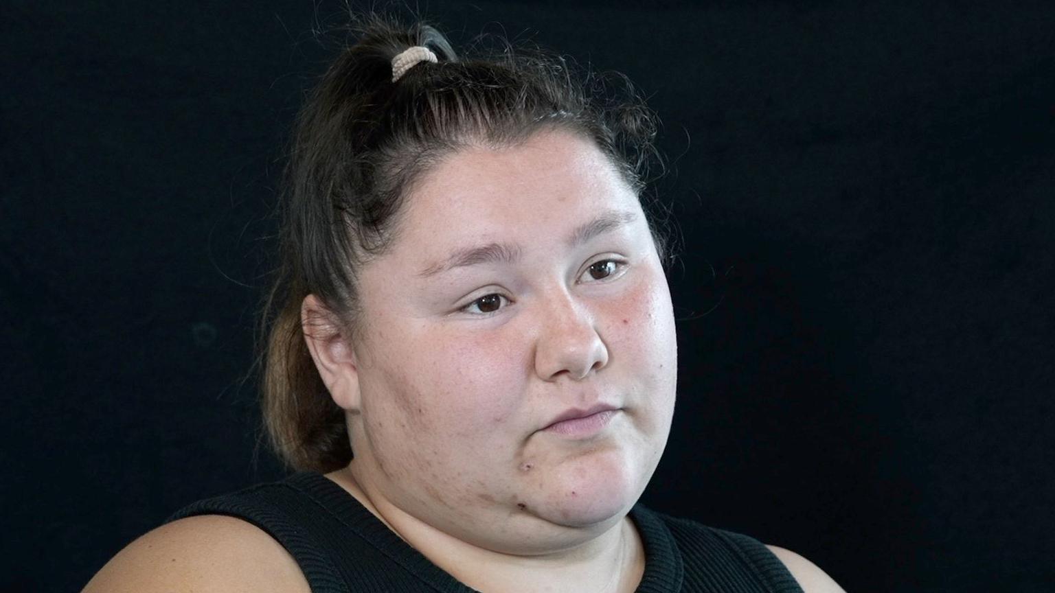 A woman with brown hair scraped back into a ponytail looks off camera in a head and shoulders image on a black background. She has brown eyes and is expressionless. She is wearing a black vest top.