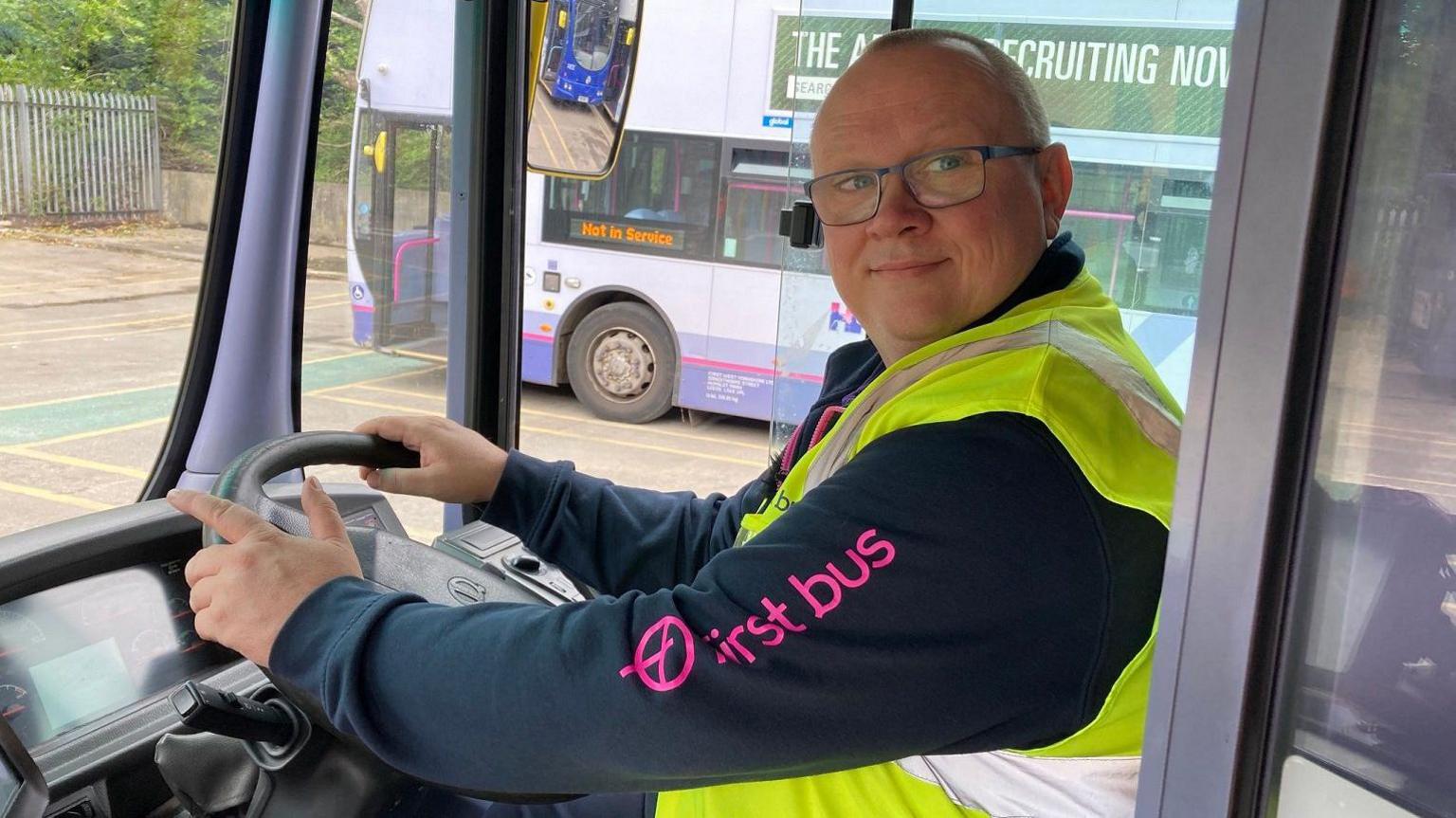 A bald man wearing a purple and blue fleece with a yellow high-vis vest over the top. He is sitting in the driver's seat of a bus holding the steering wheel.