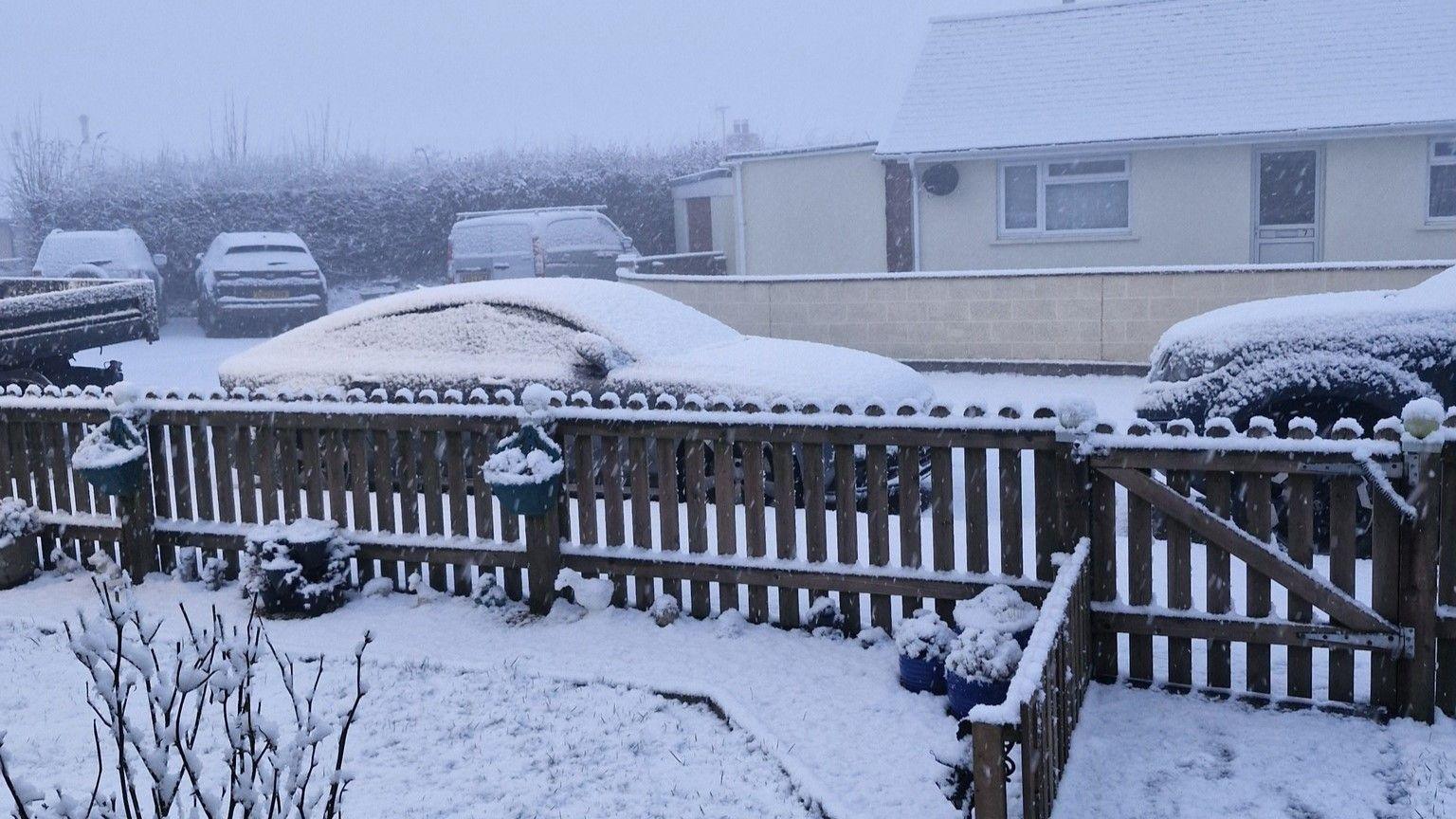 Five cars and vans parked on a residential street. Heavy snow has fallen and settled on their roofs and windscreens. There is a front garden fence in the foreground of the picture which is covered in snow. In the distance the sky is grey and foggy with low visibility. 