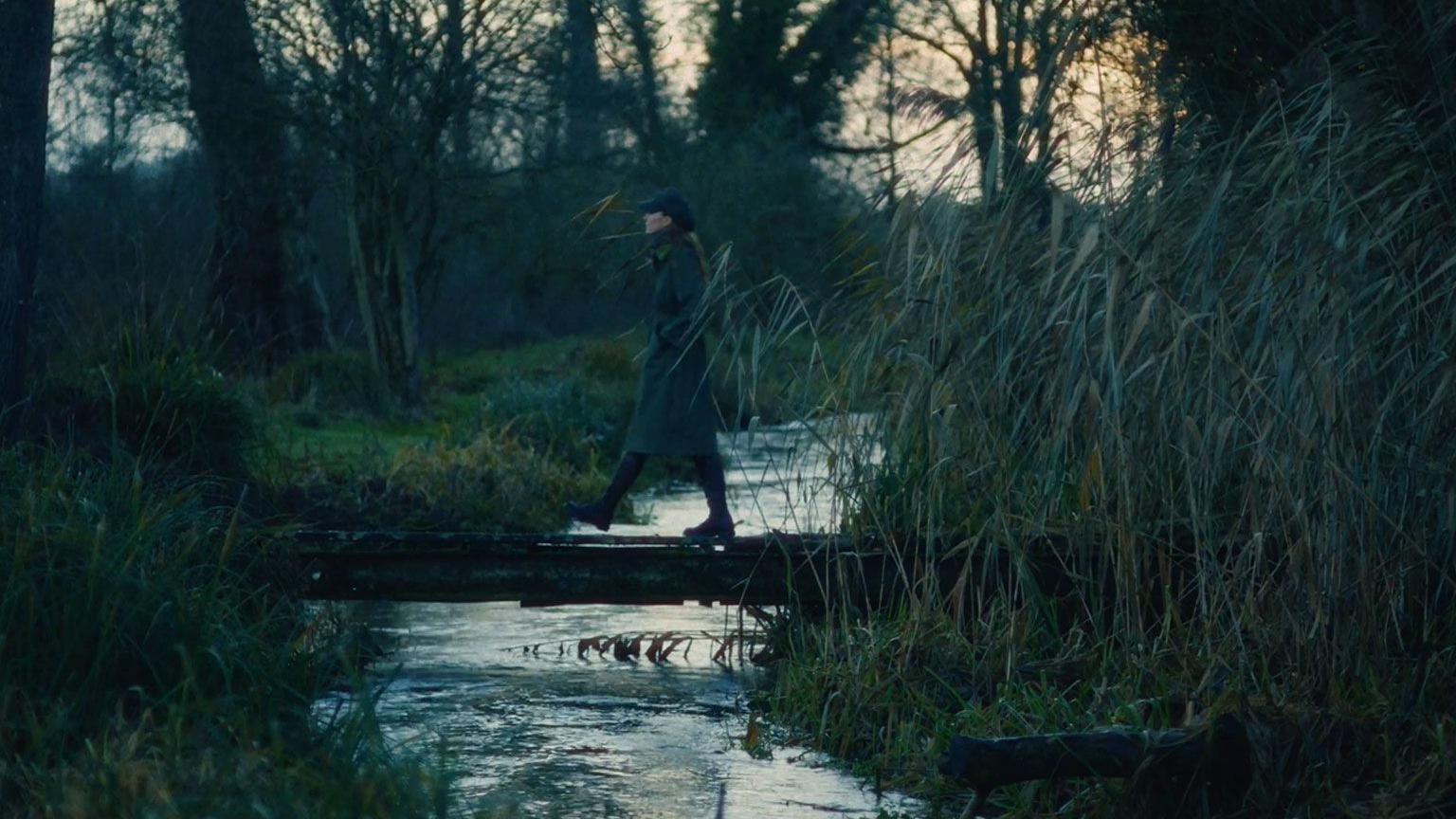 Katerina ecën mbi një urë të vogël mbi një përrua, e veshur me një pallto me gjethe të gjelbra përreth.