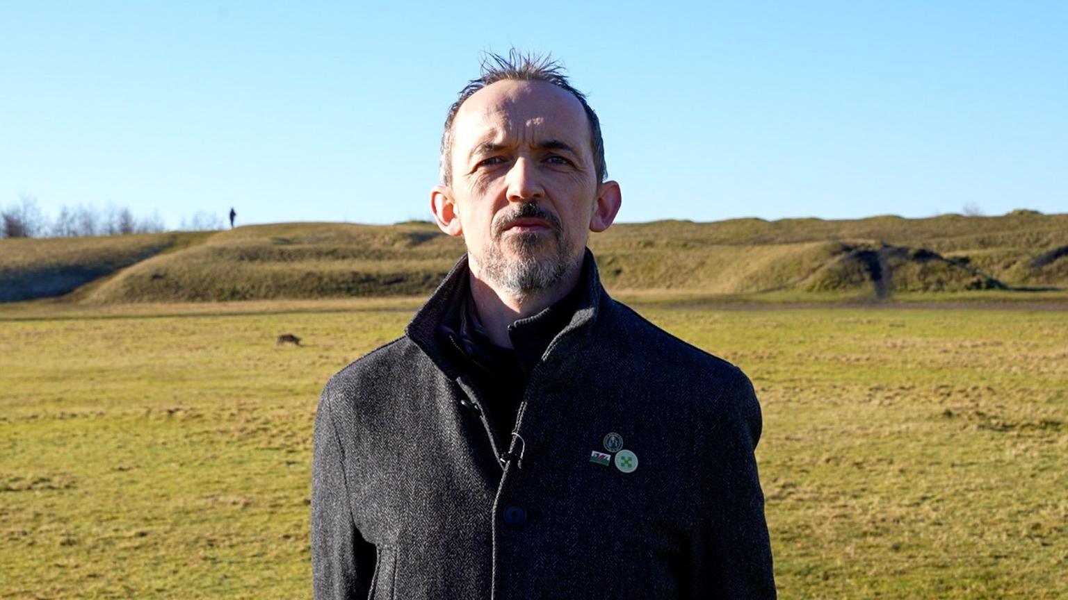 Dr Ben Curtis is looking at the camera. He is stood in a field. He is wearing a black coat and has a salt and pepper goatee.