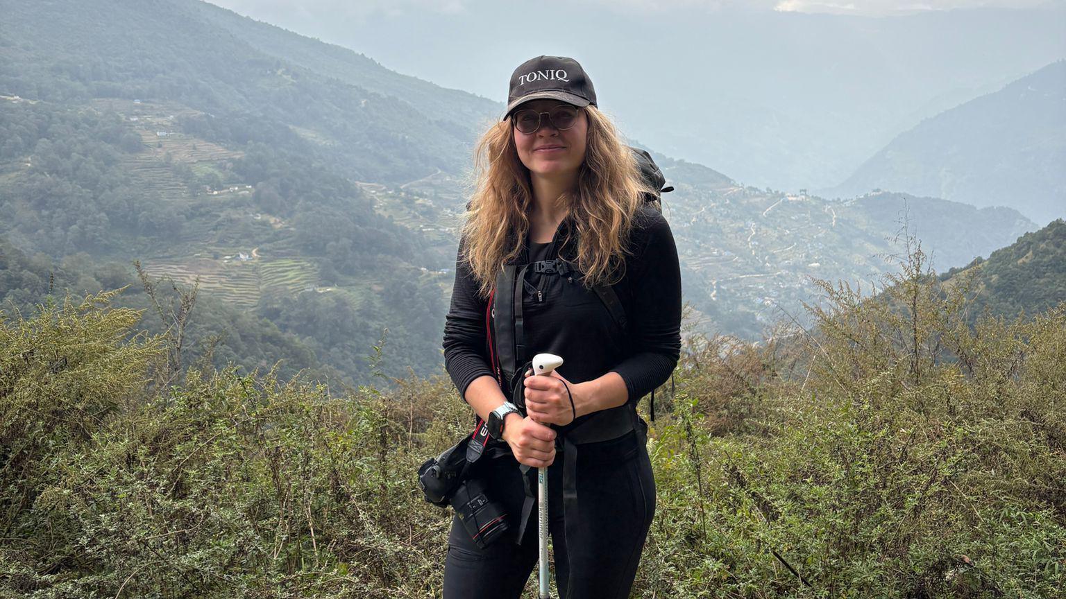 A woman is standing on a mountain holding a walking stick. She is surrounded by green plants and hills in the background. 