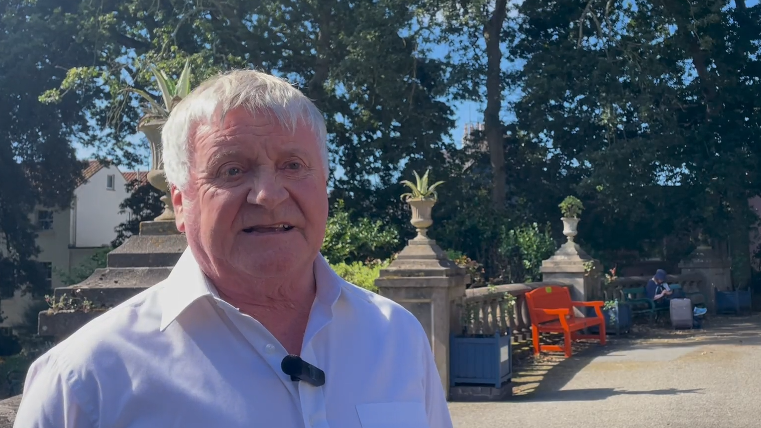 Russ Fossey, an older man with grey hair, standing in front of a stone structure with potted plants. He is wearing a white collared shirt.