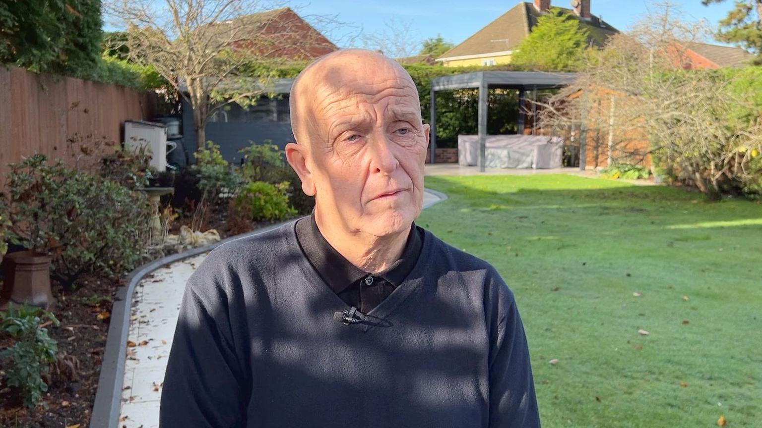 Paul Walker wears a v neck jumper and polo shirt, his expression is concerned, he is standing in a back garden, in the sunshine.
