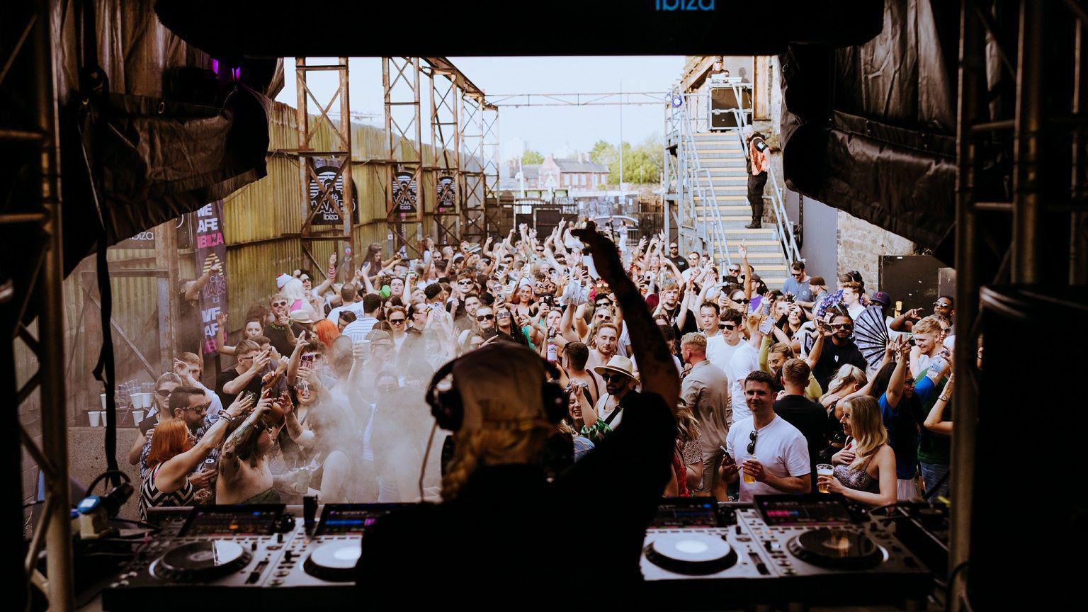 A female DJ stands behind some decks with one hand in the air and headphones on her head. She performs to a crowd of people outside of a warehouse