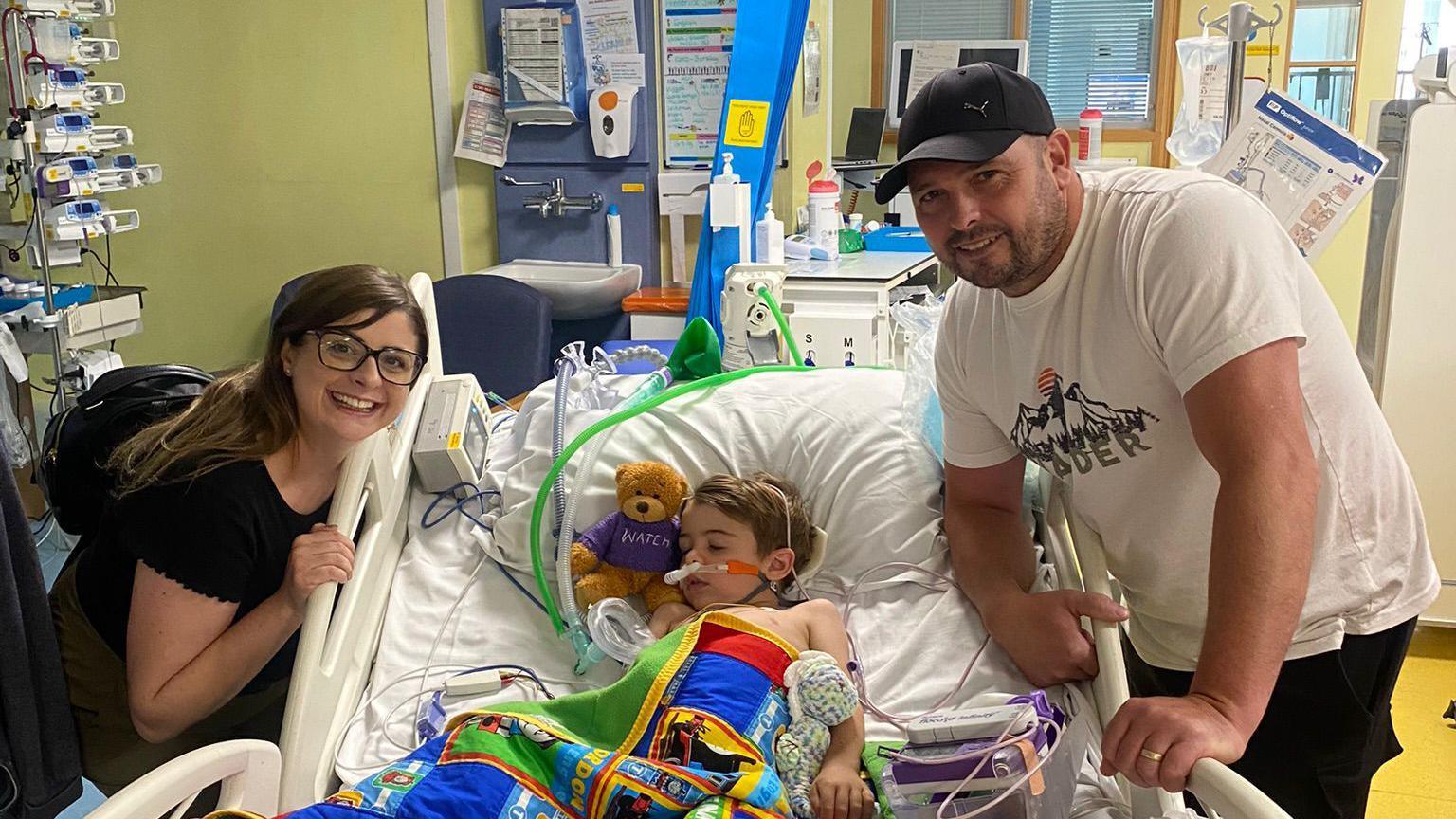 Vickie Julian and dad Steven Julian pose next to Freddie who is lying in a hospital bed. He has multiple wires attached to him and is covered with a brightly coloured blanket, in his arms is a soft toy