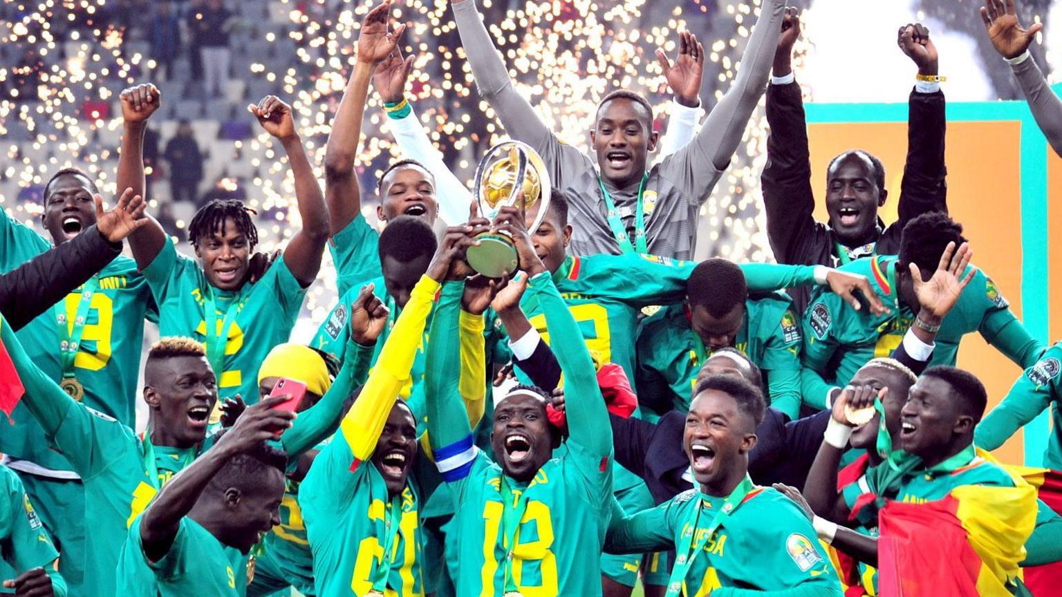 A group of Senegal players wearing green lift the African Nations Championship trophy, with flashes of fireworks seen in the background inside a stadium