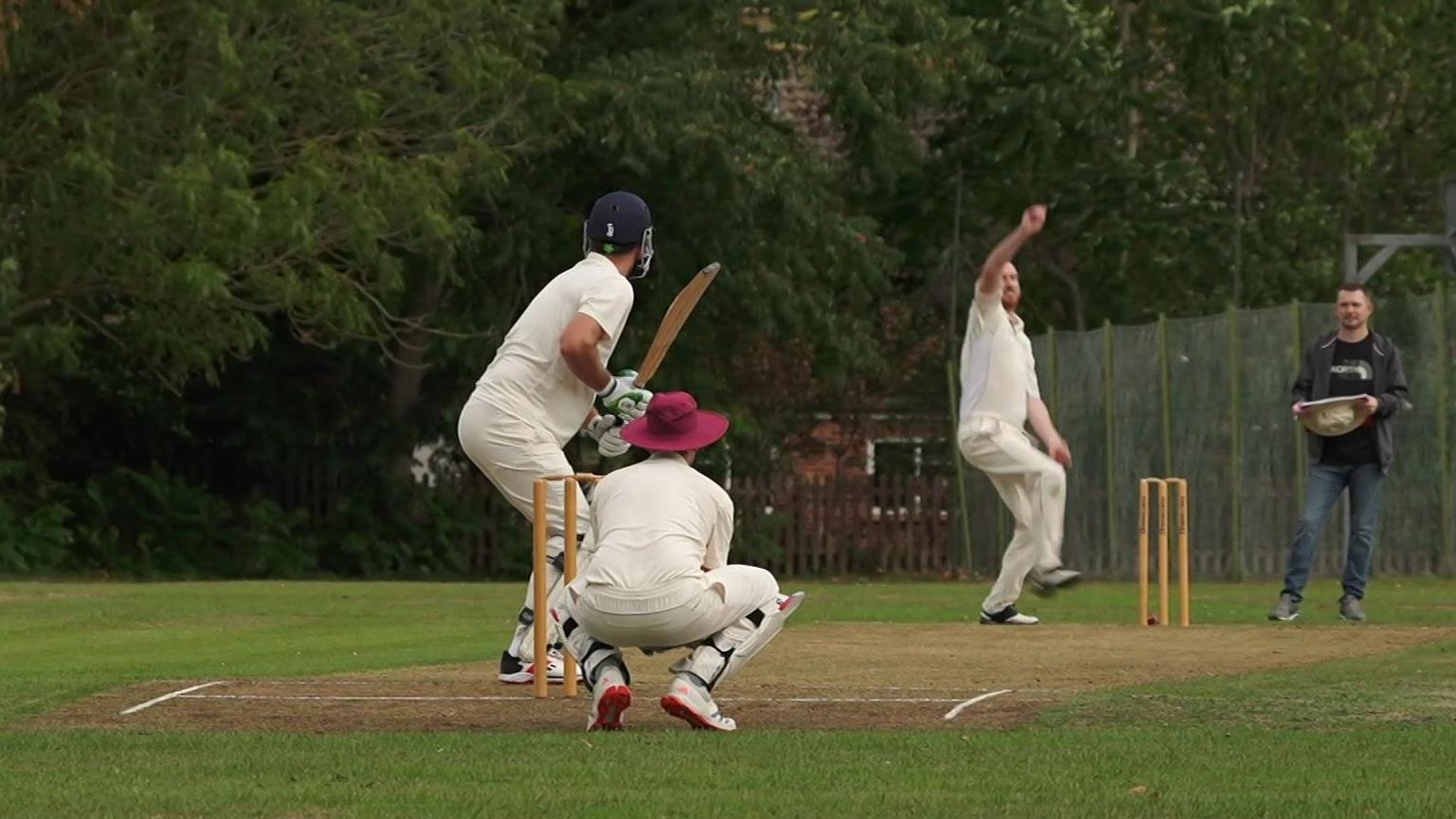 Three people are dressed in white cricket clothing playing cricket on a field. There is a person on the right watching holding a hat.