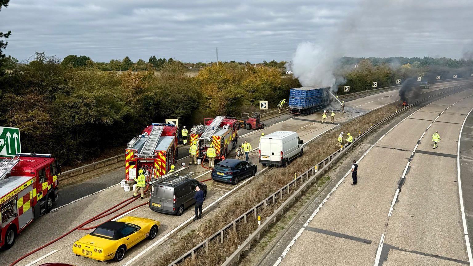 Fire engines and emergency workers on the A12. The picture is taken from above, from a bridge. There are cars at a standstill. In the background there is a lorry with a big plume of smoke coming out of it.
