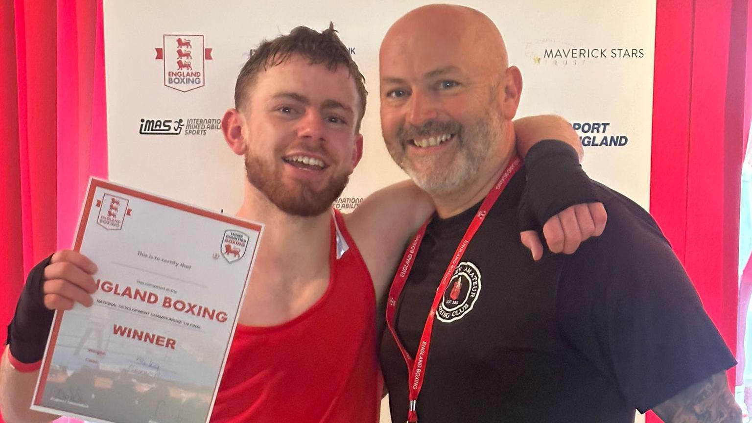 Reuben smiling at the camera, wearing a red vest and black glove holding his winning certificate.  His right arm is hung around is Dad Danny's neck, Danny is wearing a black t-shirt and smiling.