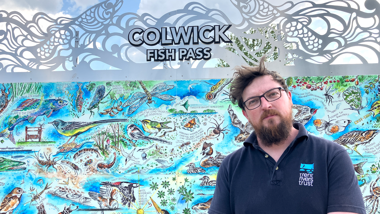 A man with a beard from the Trent Rivers Trust stands in front of a Colwick fish pass sign that is illustrated with wildlife.