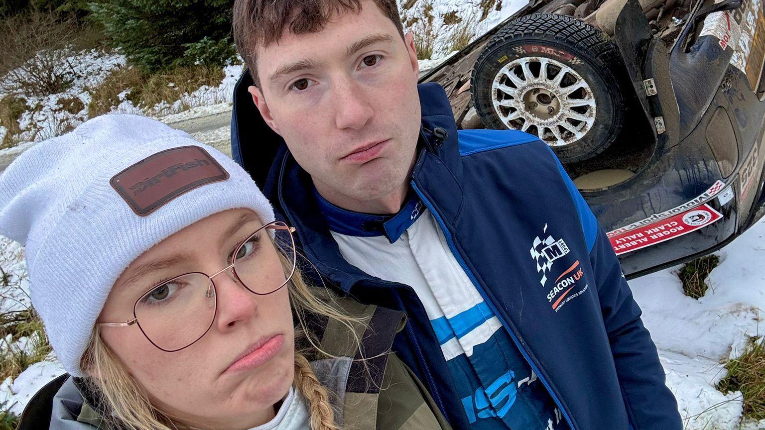 Hollie McRae and her co-driver, Jon Armstrong, with sad expressions standing in front of the upturned rally car