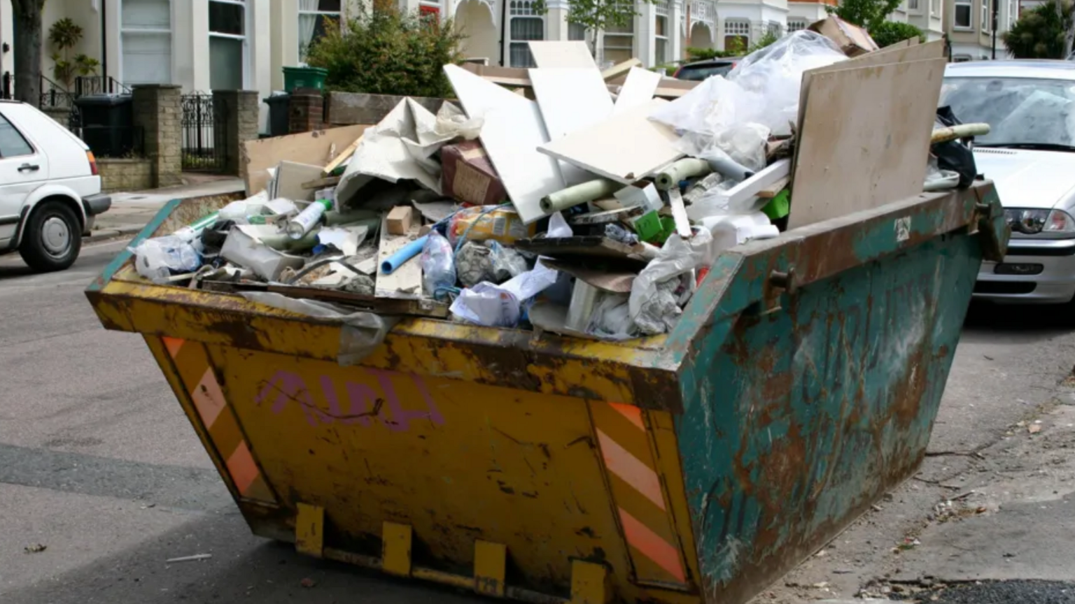 A street with a skip next to the curb. The skip is yellow on the front and blue on the sides. The skip is filled with rubbish. 