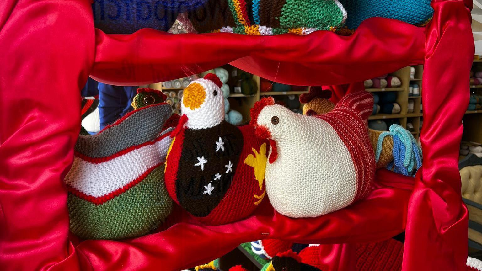 Two more of the knitted hens on display on shelf that has been dressed in a red fabric. 