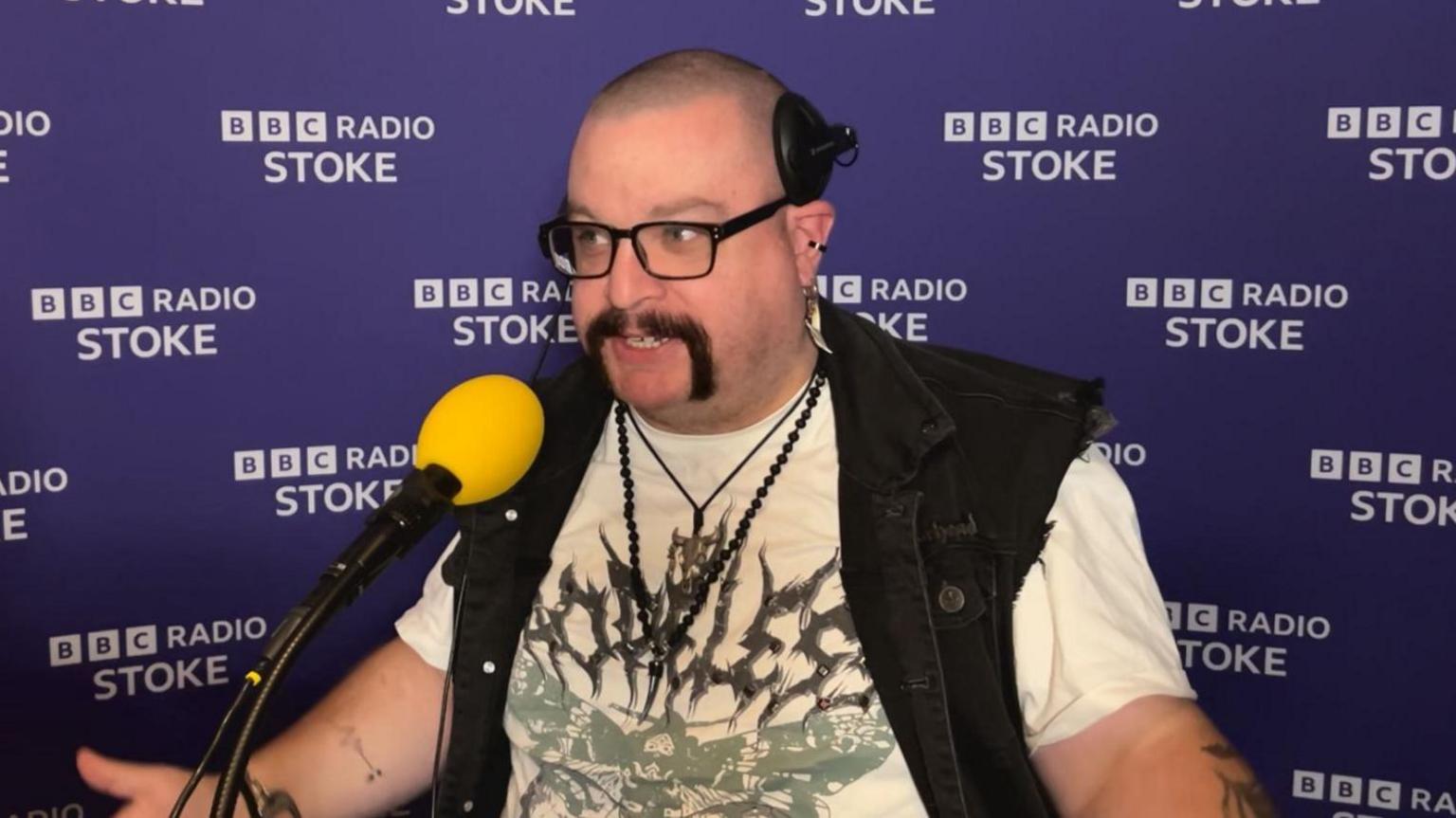 A bald man with headphones on his head, glasses, a dark denim waistcoat, a cream shirt with a butterfly logo on front and black bead necklaces, sits in front of a yellow microphone while talking.