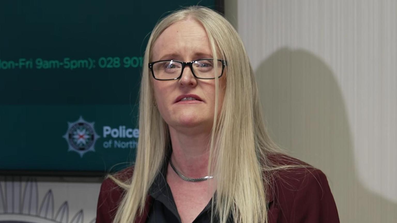 Geraldine Hanna has her mouth slightly open as she speaks at a press conference. She has long blonde hair, and silver necklace. She is wearing a wine coloured suit jacket and a black shirt.