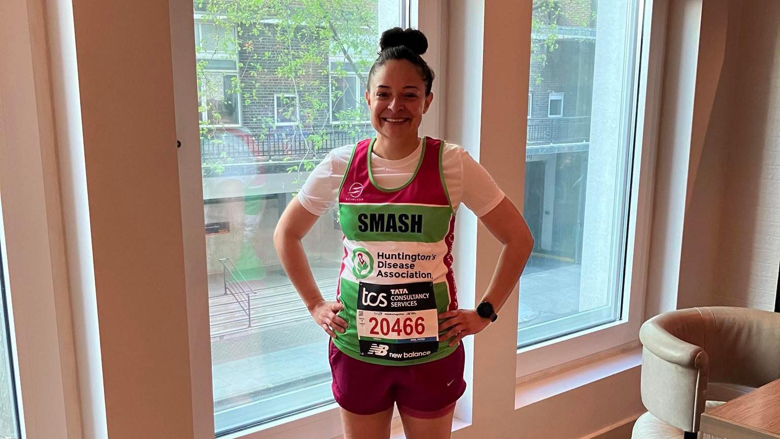 Ashley stands infront of a window by a chair smiling at the camera with her Huntington's Disease Association top on which says SMASH