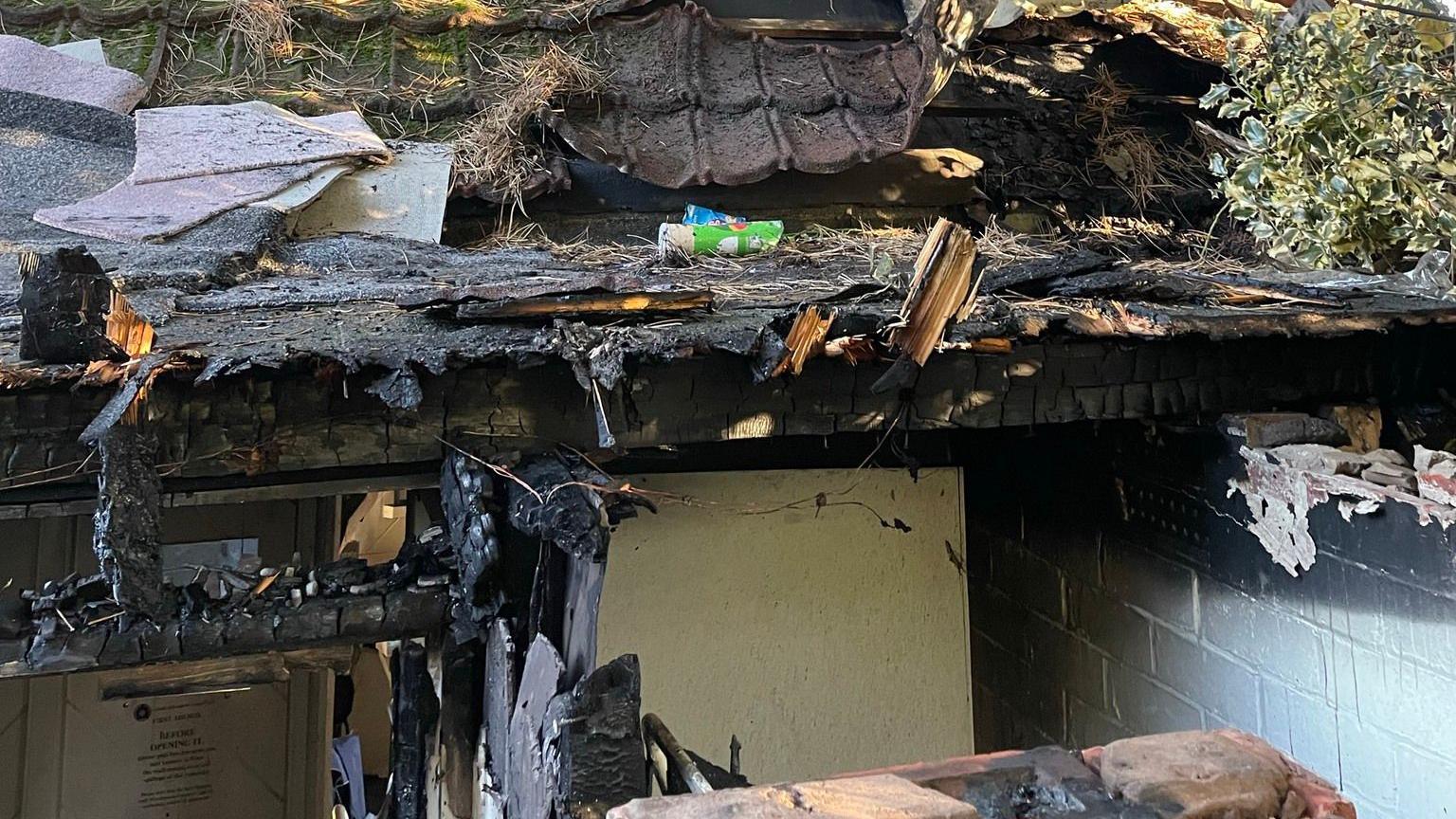 A roof surrounded by trees which has been destroyed by a fire. The edges are blackened and parts of the roof missing, exposing the rooms below.