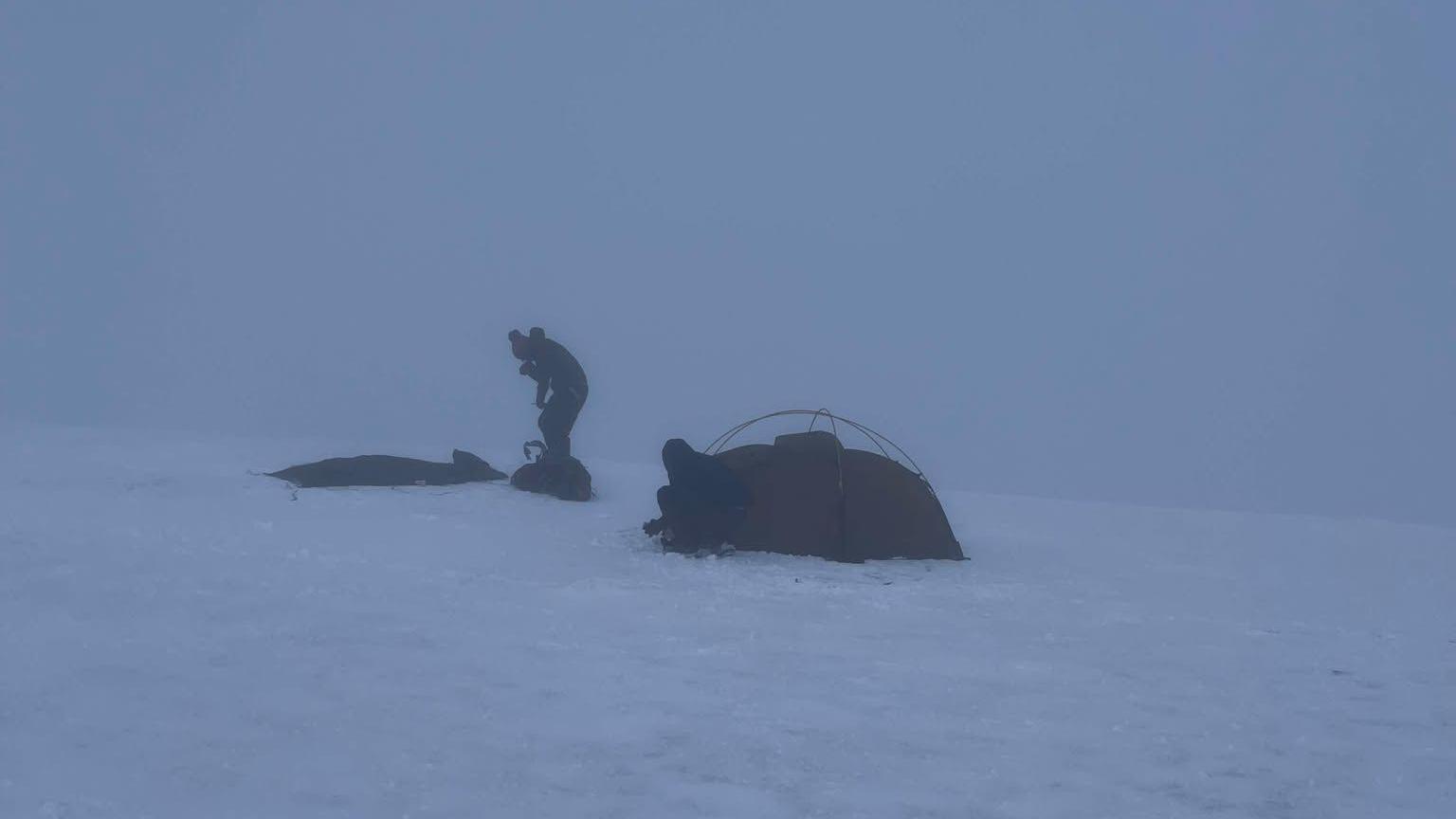 People setting up camp atop of Helvellyn in full white-out conditions. There are two people crouched trying to set up a tent.