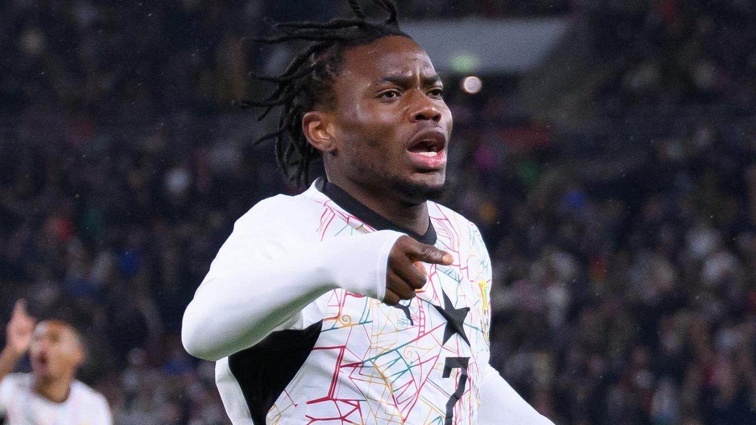 Abdul Fatawu points with his right hand and his mouth open celebrating while wearing a white Ghana shirt after scoring against Germany in a friendly.