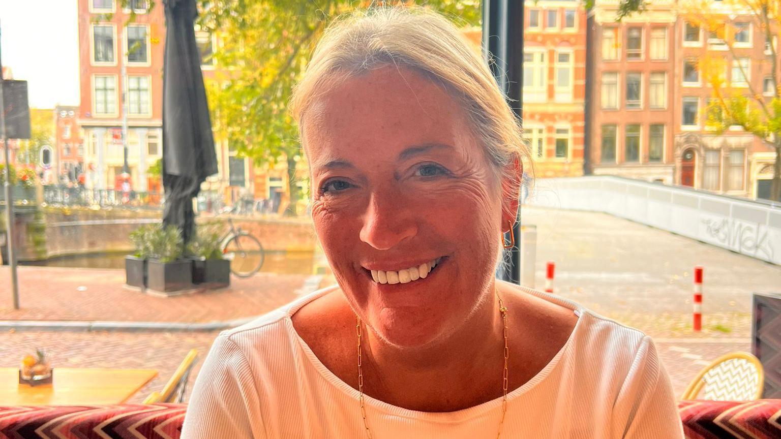 A woman with light blonde hair wearing a white top and a gold necklace, smiling at the camera. A cobbled street and a row of buildings is in soft focus behind her.