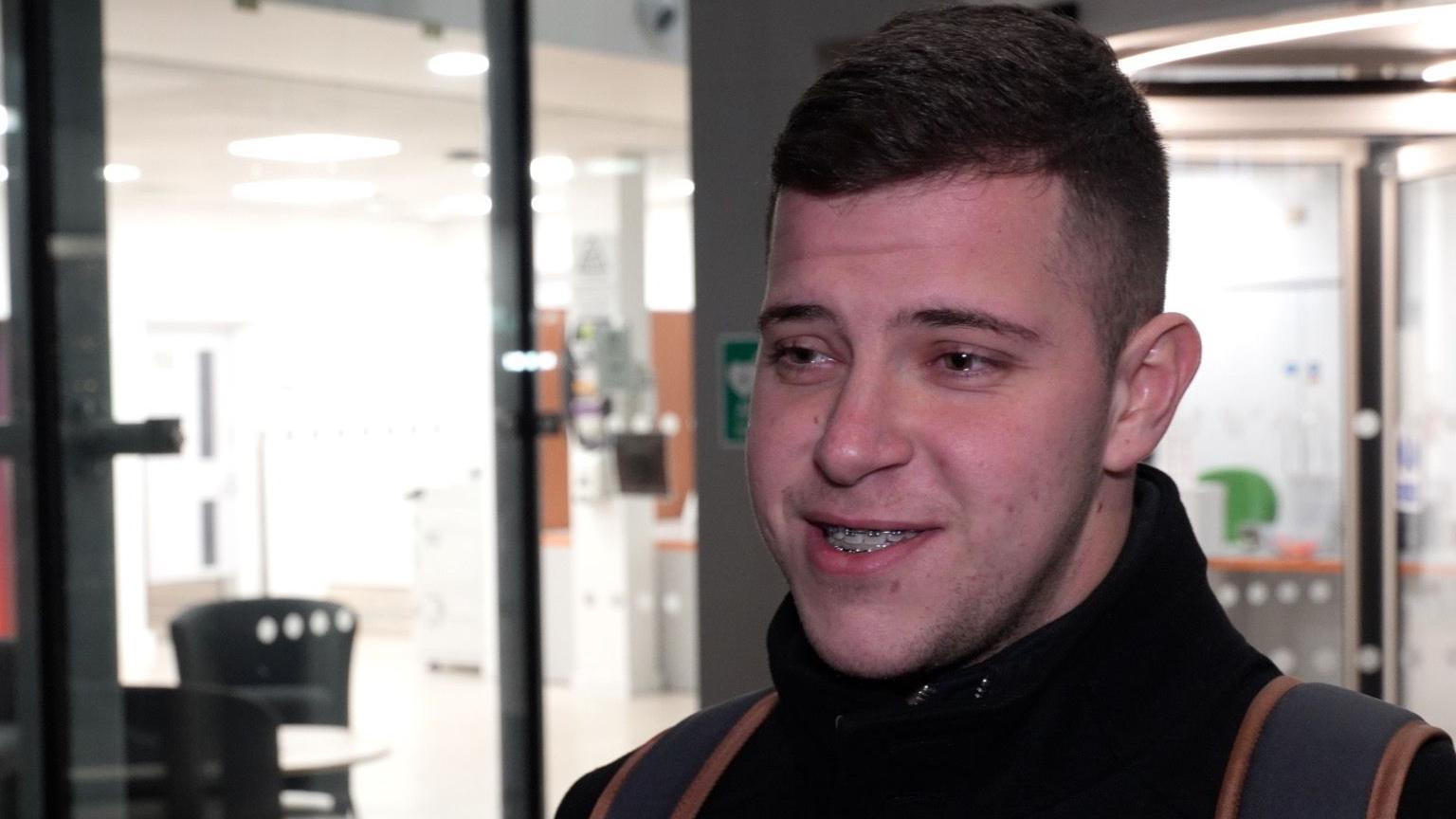 Preslav Hristov looks to the left of the image as he is talking. He has short dark hair, braces, and wears a black coat with a large collar. A grey and orange rucksack is on his back. He stands in front of a university building with large windows. 