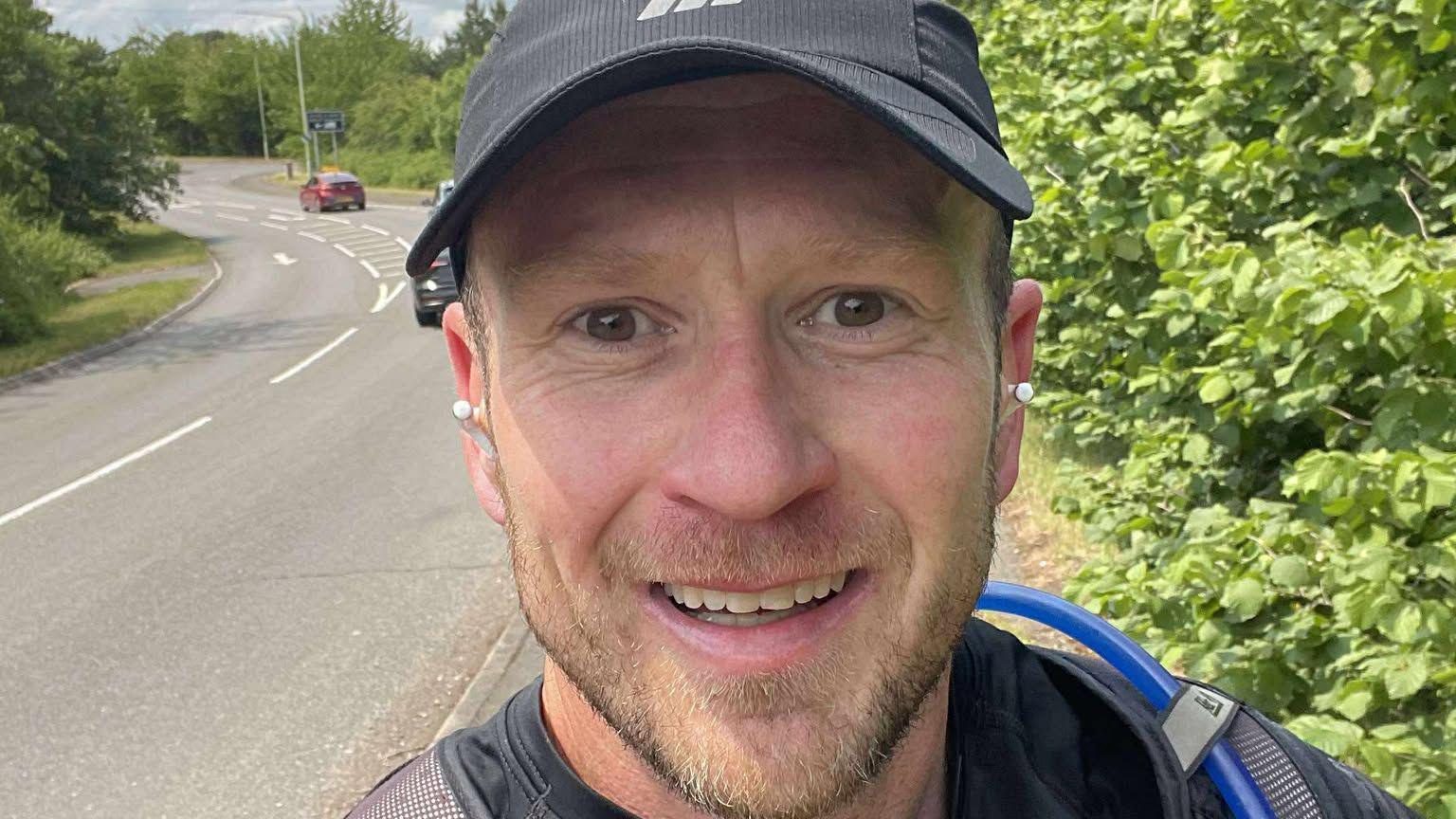 Mike Jones taking a selfie alongside a road with shrubs to the right and the road on the left.