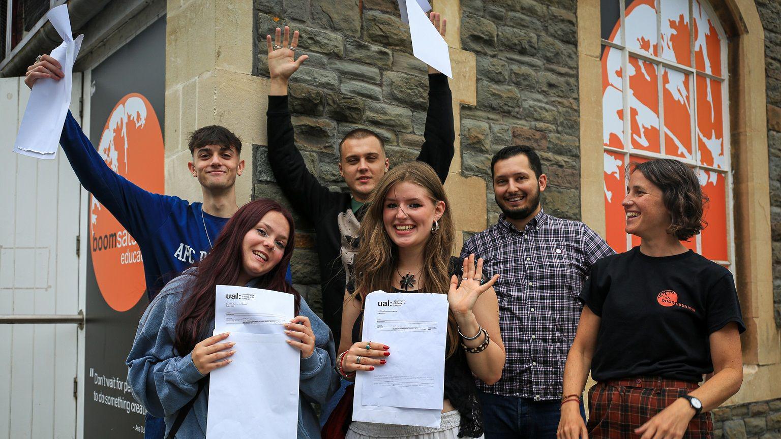 A group of A-Level students from Bristol education provider Boomsatsuma stand outside a brick building. They are celebrating and holding up their exam result papers.