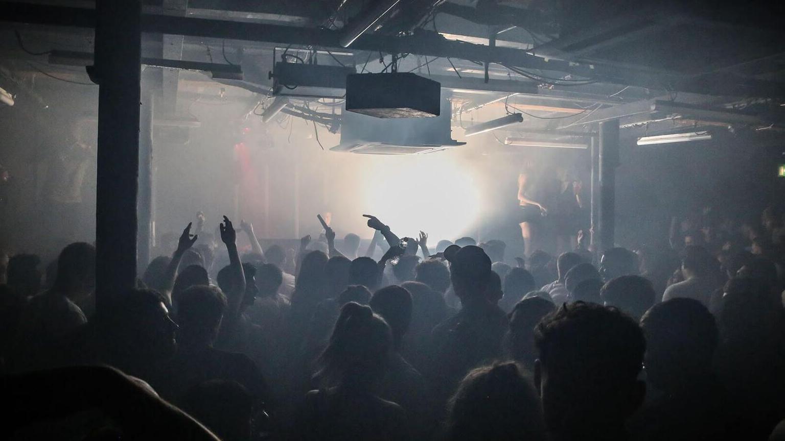 Silhouetted figures are shown dancing with arms aloft in front of a bright white light inbetween pillars in Sankeys Soap in Manchester