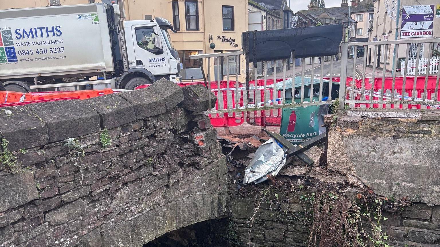 A small stone bridge that is crumbling on one side, surrounded by an orange barricade and twisted railings. A waste management lorry can be seen on the road behind as can a number of businesses, including a cafe and a card shop.