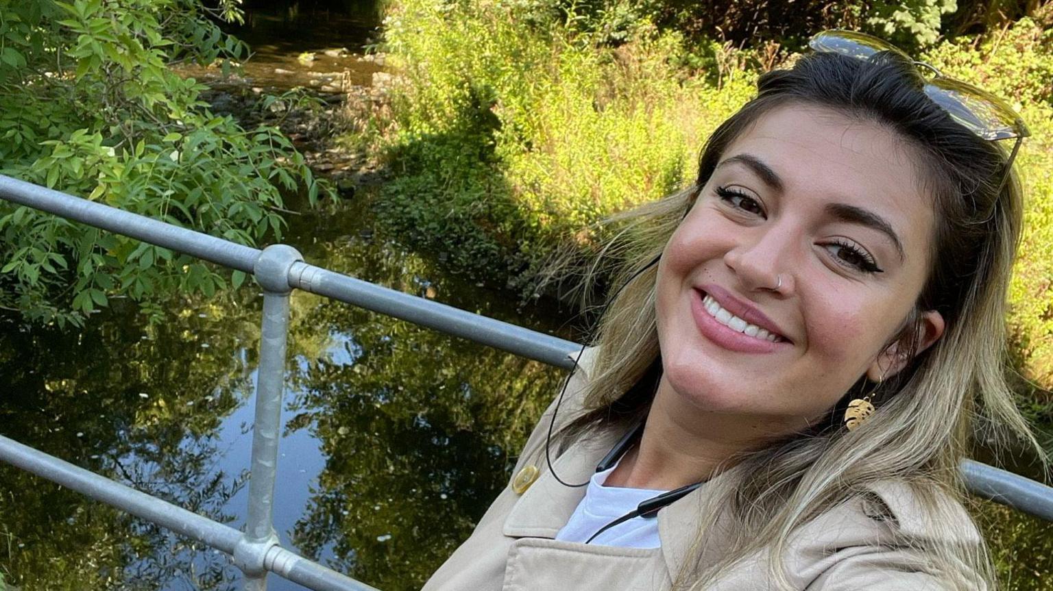 Selfie of Ruby Lake, a young woman with brown hair, smiling next to a body of water and sunglasses on her head.