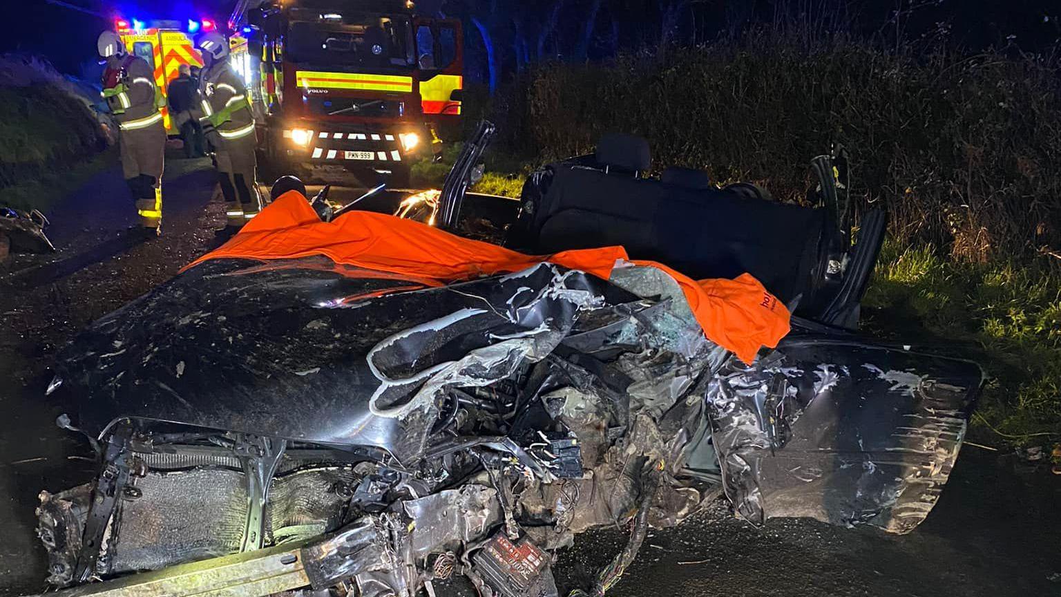 The badly damaged wreck of a dark coloured car that had had its roof cut off. It has a bright orange blanket draped over part of it. A fire engine with bright lights and two firefighters can be seen the in the background.