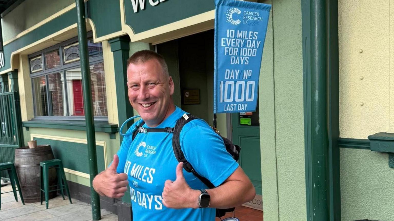 David Ansell smiling into the camera with both of his thumbs up by his chest. He is wearing a bright blue top with the Cancer Research UK logo and '10 miles for 1,000 days' written on it. He is wearing a black backpack which is strapped across his chest which has a blue flag hanging off the back of it. The flag says the same as the T-shirt, but also reads 'Day No 1,000 last day'.