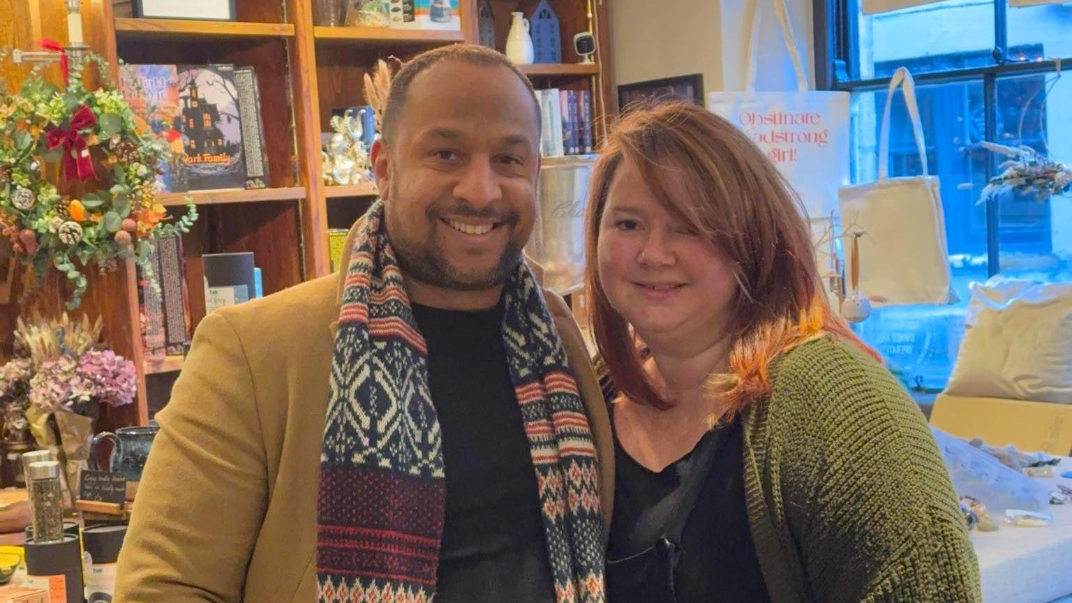 A man and a woman stand smiling inside a shop. Shelves behind the counter are full of books, tote bags and bunches of flowers.