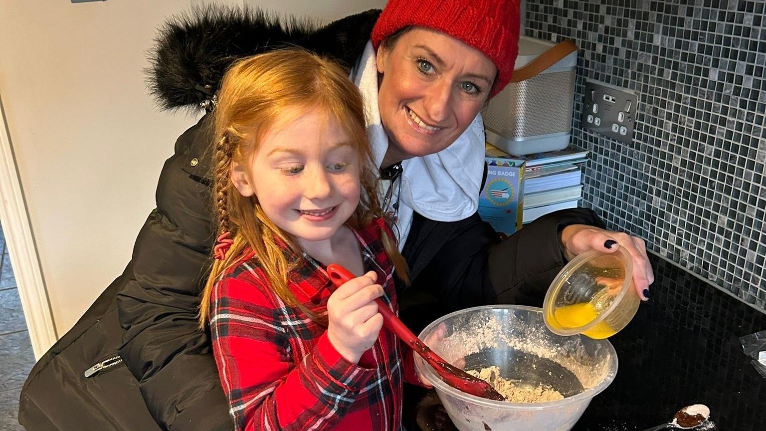 Susannah is wearing a red wooly hat and fluffy hooded coat and is smiling at the camera. She is baking a cake with her daughter who is stirring a red spoon in a bowl. Her daughter has long ginger hair and is wearing a red and black checked shirt.