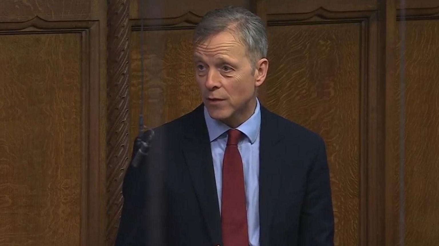 A grey-haired man wearing a suit speaking in Parliament with a wooden panelled wall behind him.