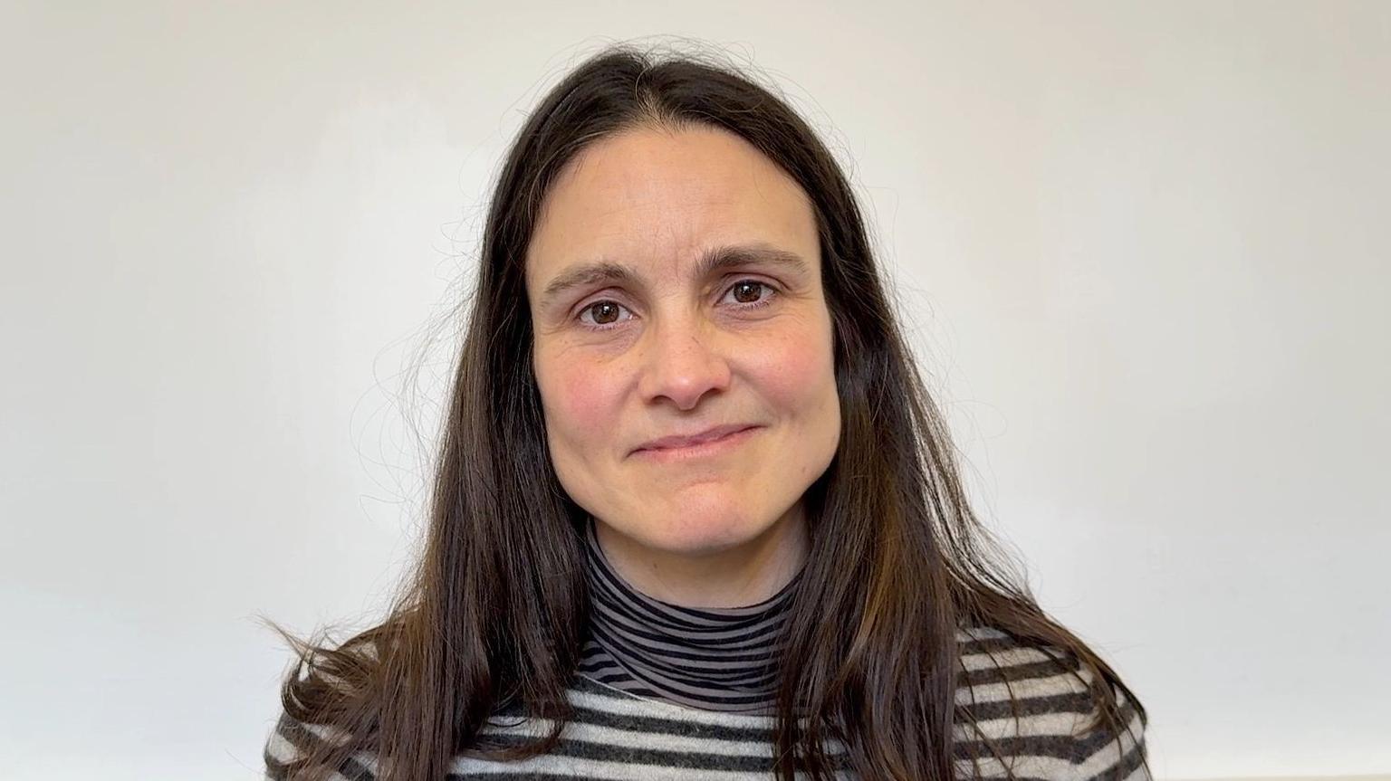 Dr Karen Mansfield from Oxford Internet Institute. She is standing in front of a cream background wearing a black and white striped polo shirt. Her long dark brown hair is over her shoulders. 