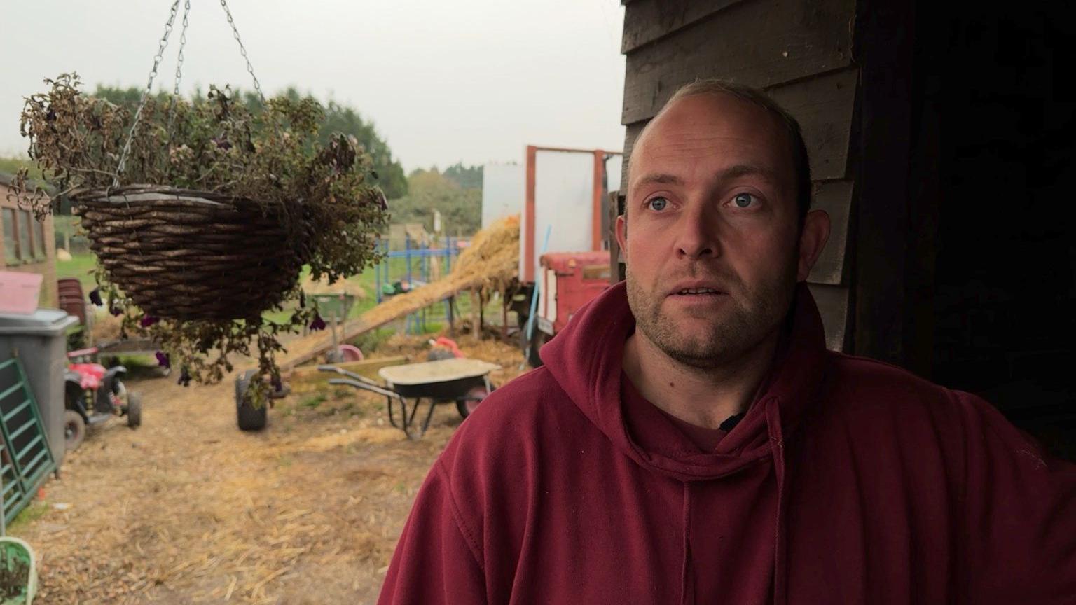 Kieran Loder stands in a section of farmyard, there is a hanging flower basket nearby and gardening tools behind him to the left and the outline of a wooden panelled shed on his right. Kieran wears a deep red hoodie and looks slightly off camera.