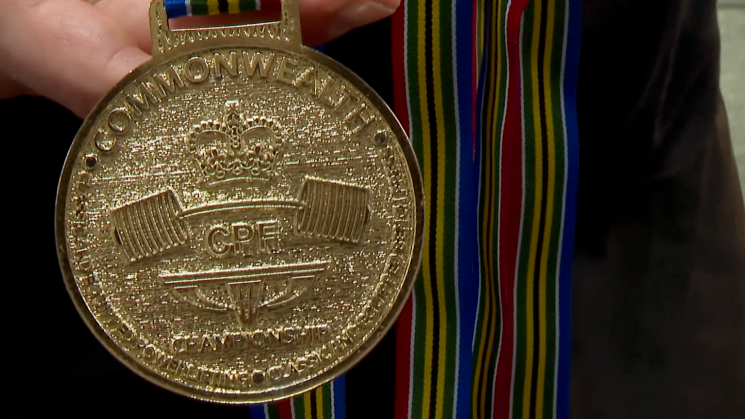 Commonwealth Powerlifting Federation Gold Medal for Deadlift being held by Lucas Williams. The gold coloured medal depicts a bending barbell with several weights on each side of the bar, with the letter CPF in the centre and Commonwealth at the top and Championship at the bottom. The medal is held against multicoloured medal ribbons