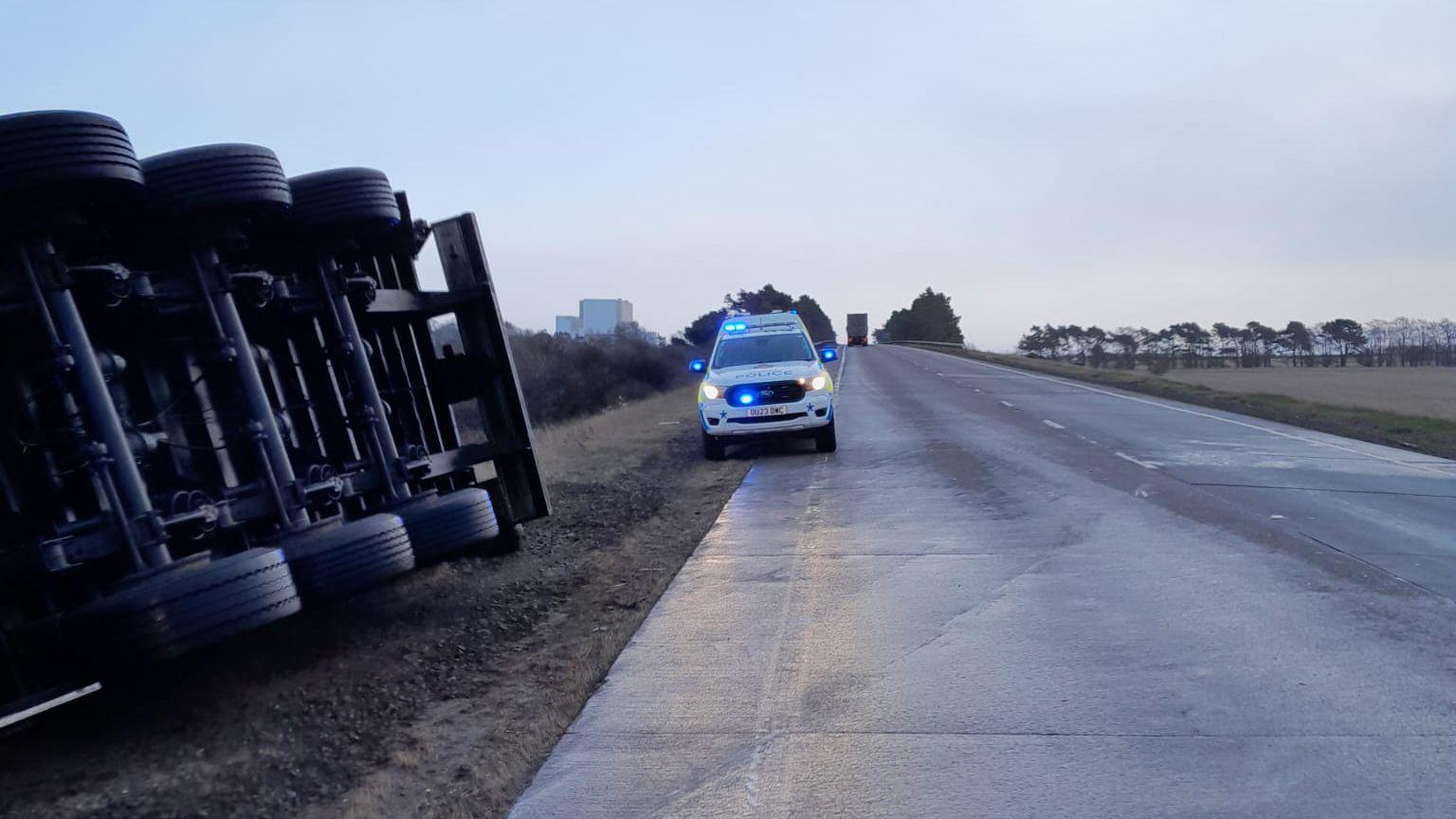 An overturned lorry by the side of the road on the A1 in East Lothian. Only the wheels of the lorry are visible. There is a police car with its lights on in the middle of the shot.