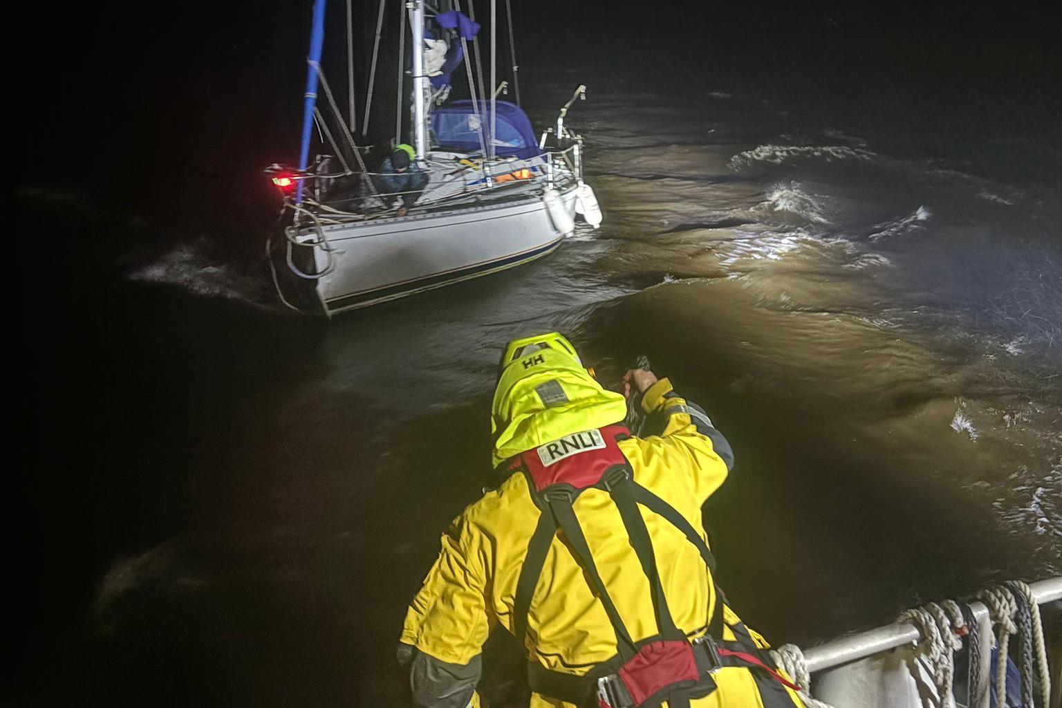 A rescuer in a full yellow high vis out fit in the foreground reaches out towards a yacht in dark stormy waters