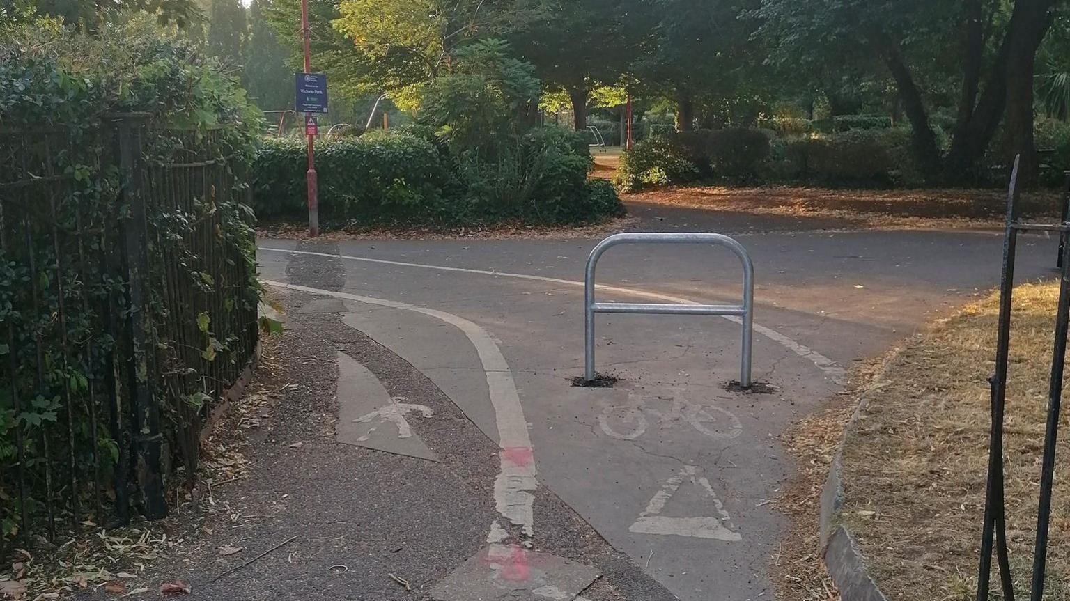 A metal bike barrier is in the middle of a cycle path at the entrance to a park, obstructing cyclist's way in. Around the path are various patches of grass and hedges