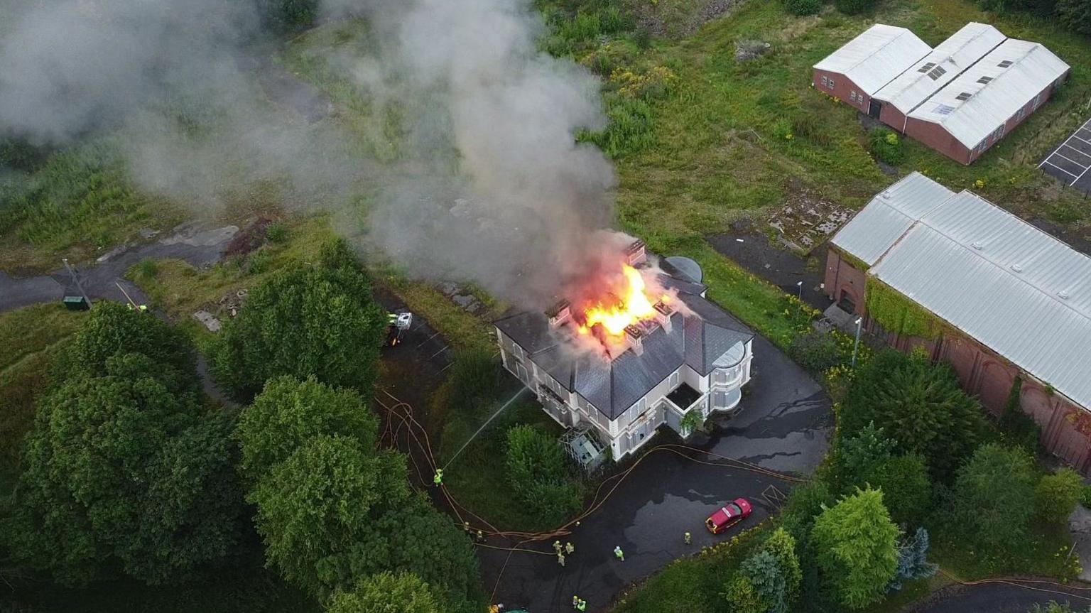An aerial view of the fire at Duncreggan House. There are flames and smoke coming from the roof of the large two-storey building. A number of other large buildings are nearby and there are trees and grassland around the burning house.
