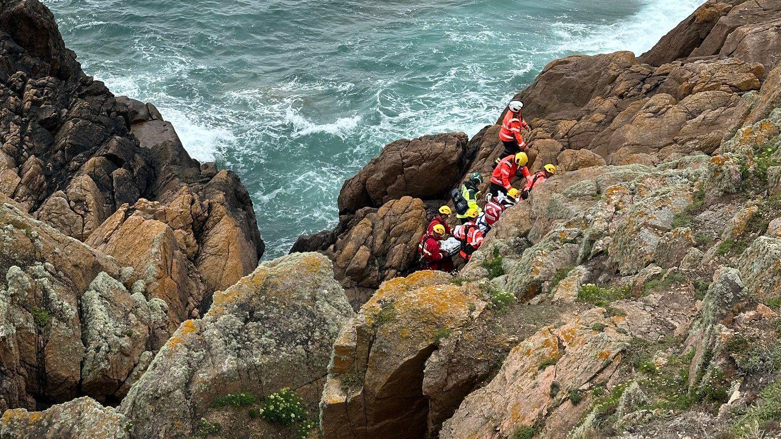 Nine emergency service workers wearing high-vis uniform and helmets are grouped together in a gap between some rocks. The cliffside is steep and rocky. The sea is a short distance from the group and it is choppy.