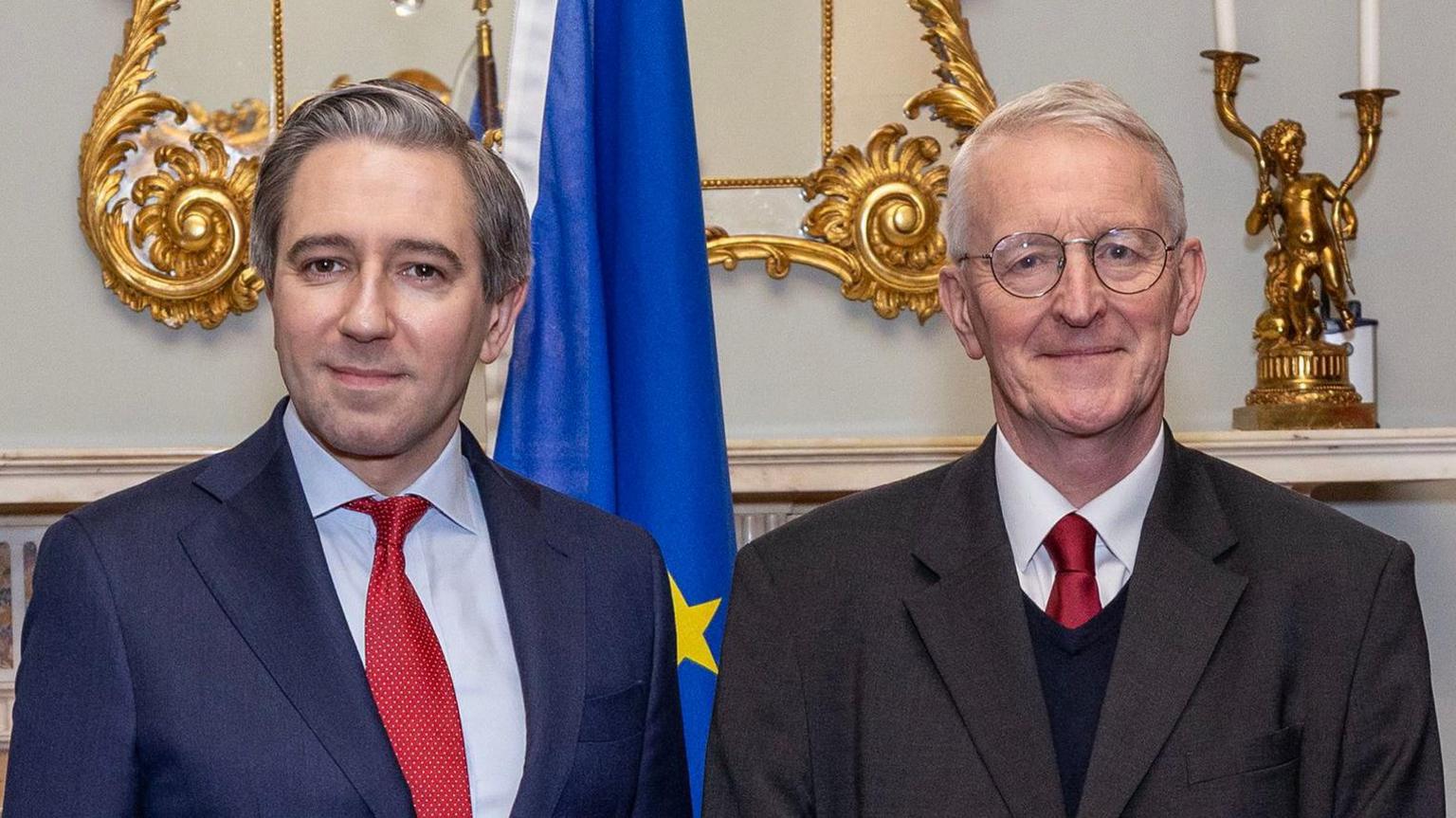 Hilary Benn and Simon Harris standing beside each other. Benn is wearing glasses, a black suit, white shirt and red tie. Harris, is wearing a navy suit, burgundy tie and white shirt. There is an EU flag behind them and a golden framed mirror behind and a golden candlestick holder .