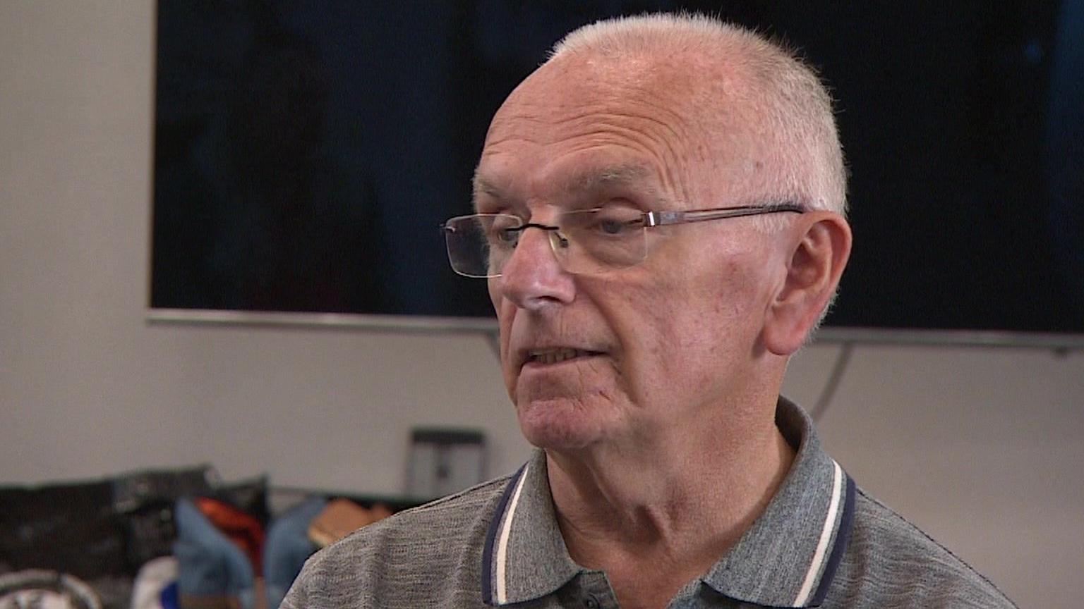 Peter Cole sits on a chair while talking to another person off-camera. He is wearing a grey polo shirt and has glasses on.