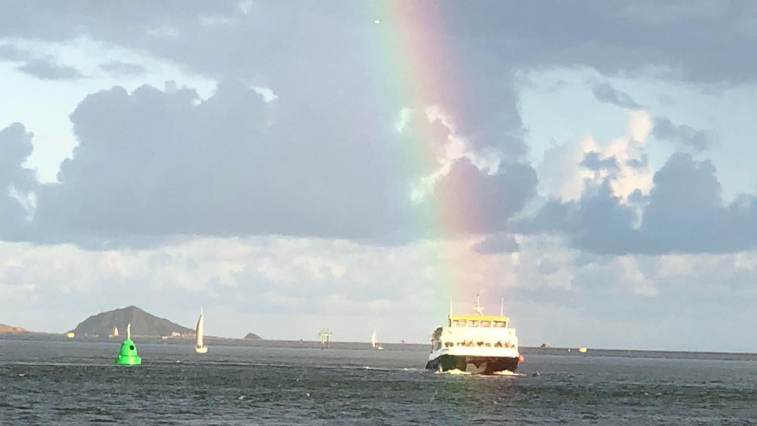 A rainbow appears to be shining on a boat in the sea near Plymouth. Several other boats and a green buoy are also on the water.