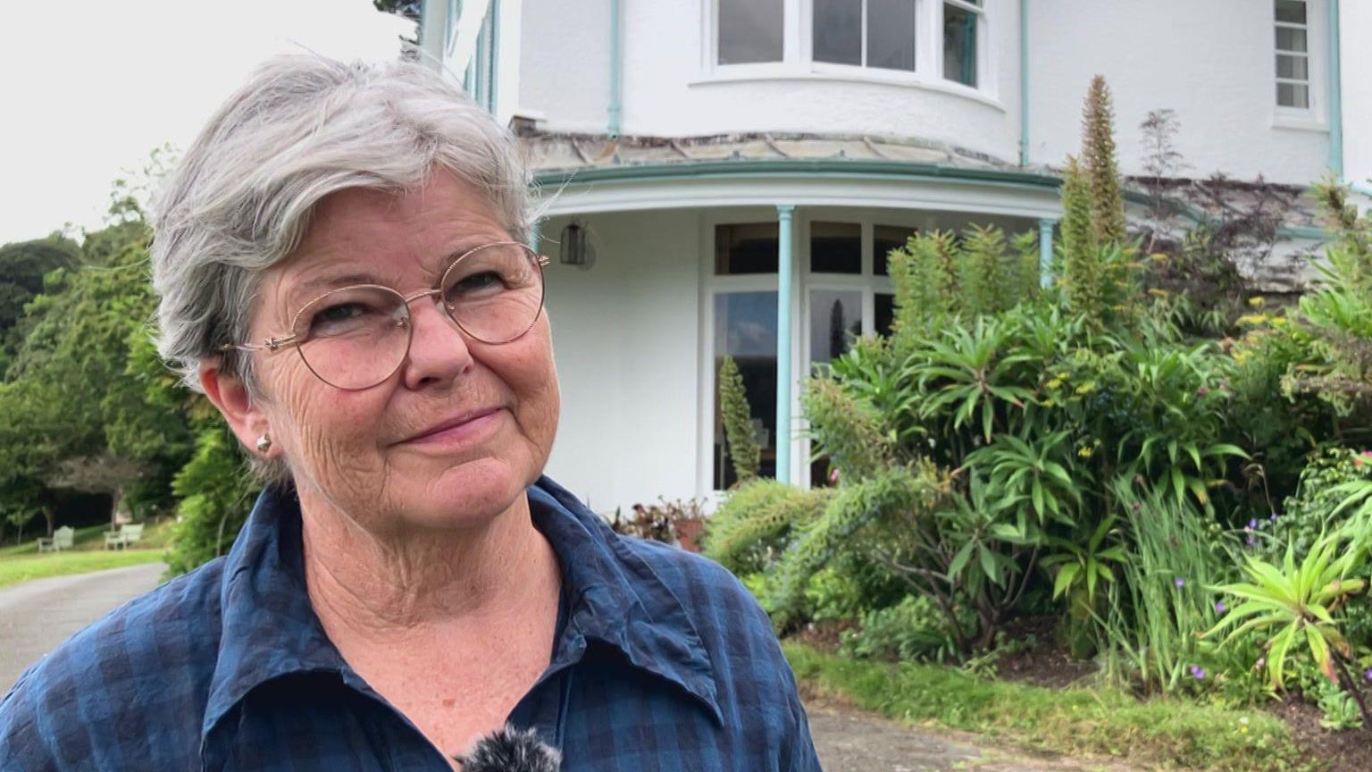 Kathy Koo smiling at the camera. She has round glasses and short hair. She is wearing a blue and black checked shirt. Behind her is the white house with green plants either side.