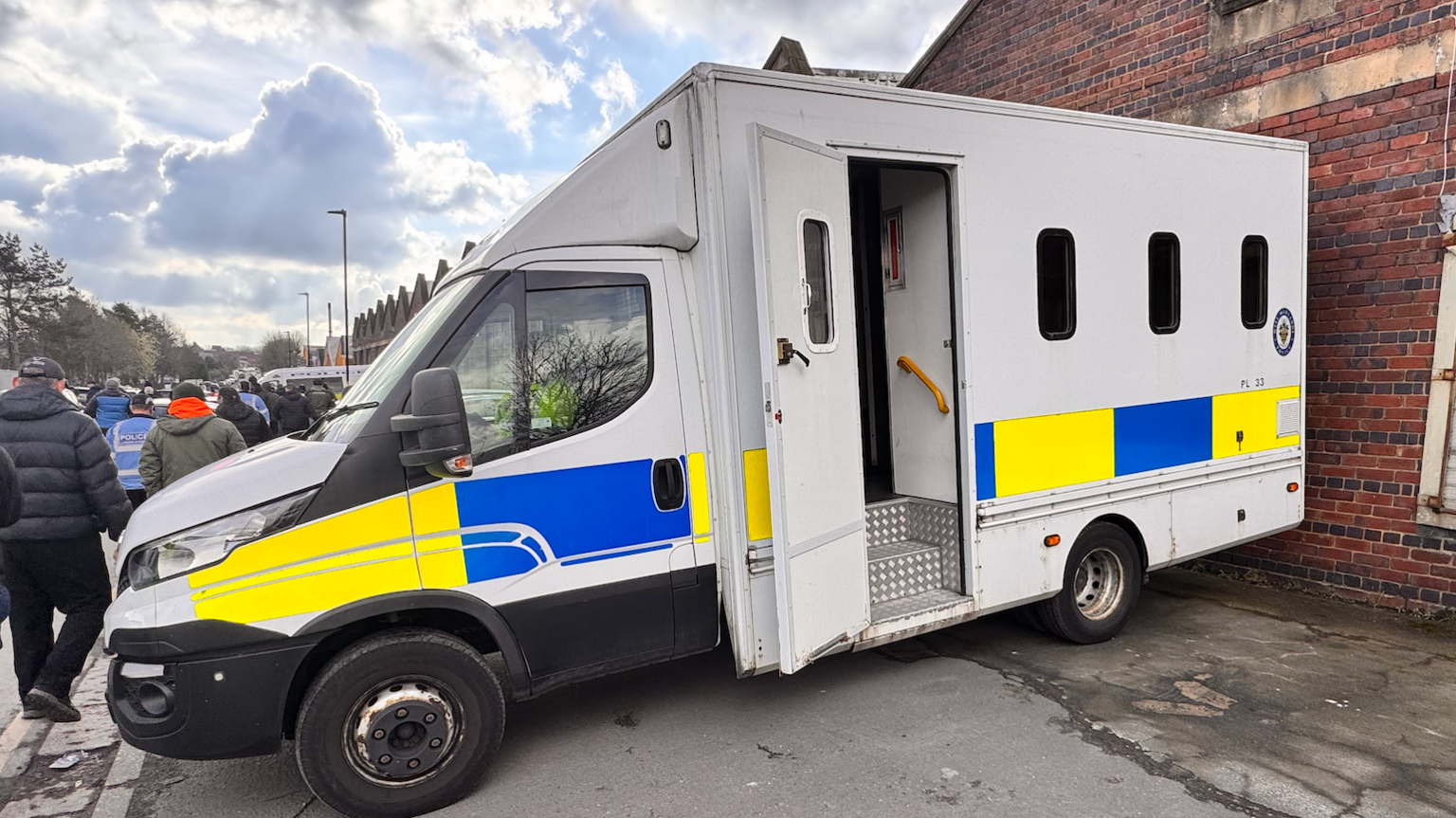 A police van parked up against a brick building. People can be seen walking on the street alongside it