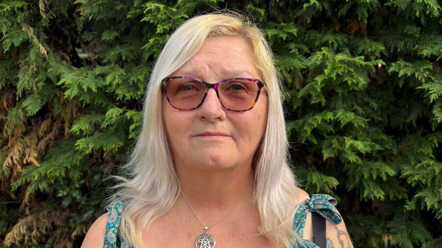A woman with long blonde hair to her shoulders stands in front of some green trees. She wears sunglasses and a metal star-shaped necklace on a chain. The top of her dress straps, green with a paisley pattern, can just be seen
